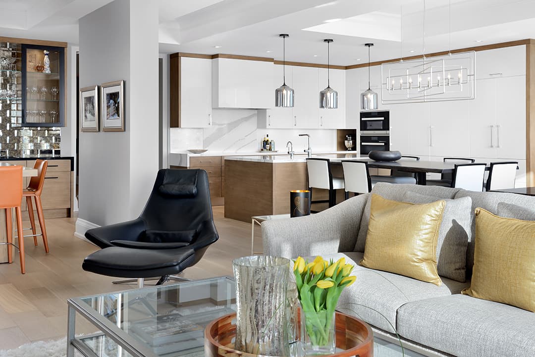 A modern open concept living space. A black leather lounge chair and a grey sofa sit in the foreground. A white kitchen with flat panel cabinets and wood accents fills the background
