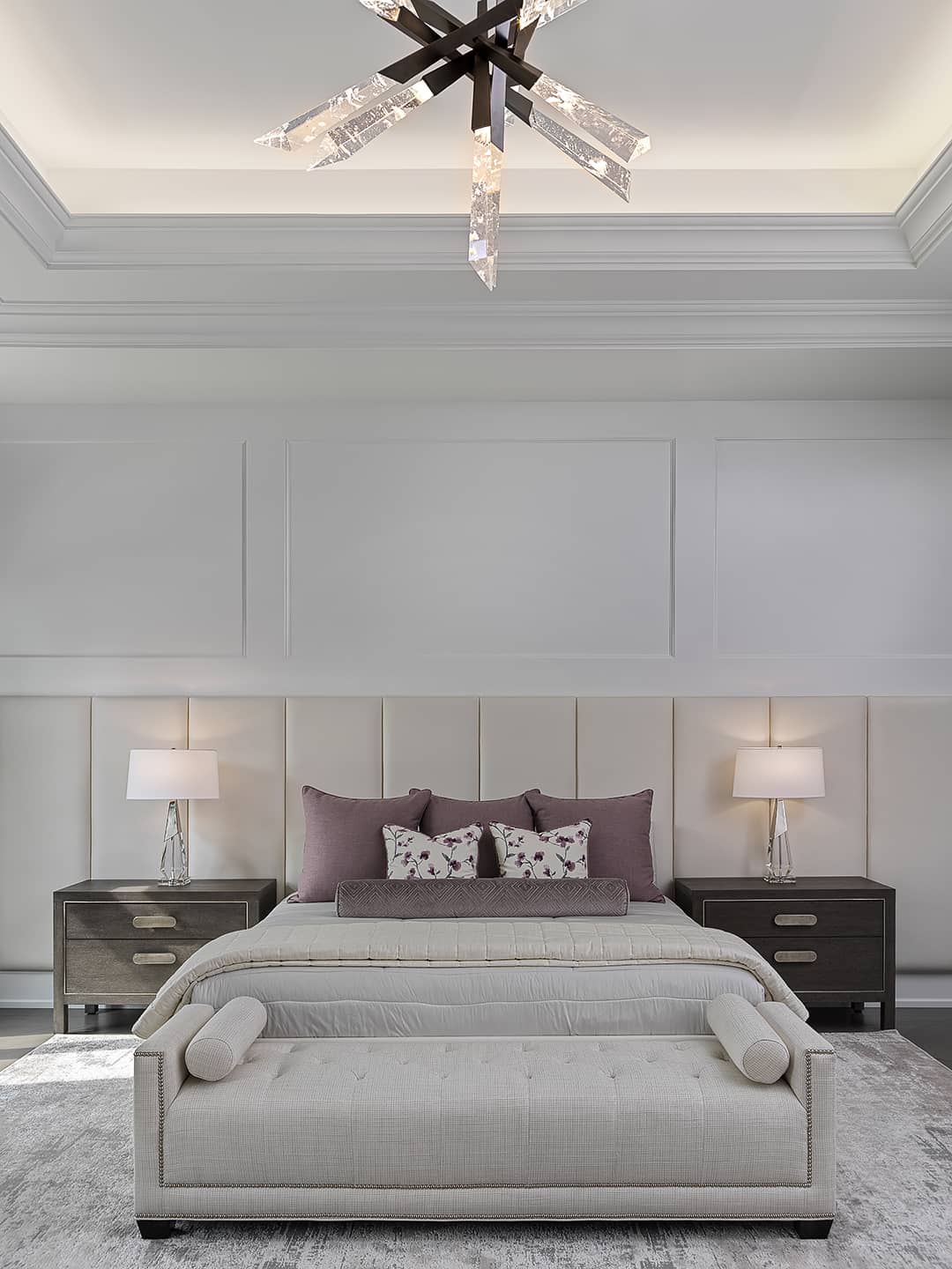 Modern bedroom with upholstered headboard wall, neutral tones, accent pillows in mauve, and crystal starburst chandelier.