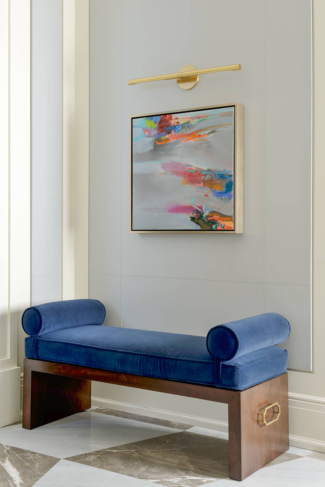 A wooden bench with blue velvet upholstery and bolster pillows, placed under a vibrant abstract painting illuminated by a brass picture light.