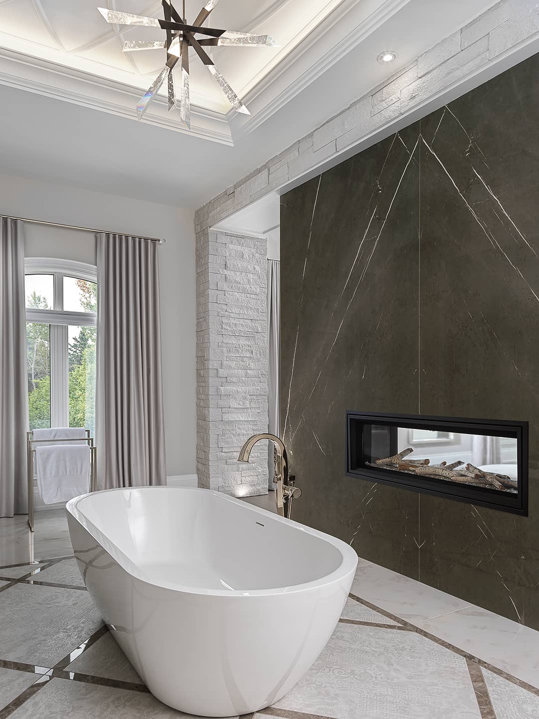 Elegant ensuite bathroom with freestanding white tub, double-sided fireplace, coffered ceiling, and crystal chandelier.