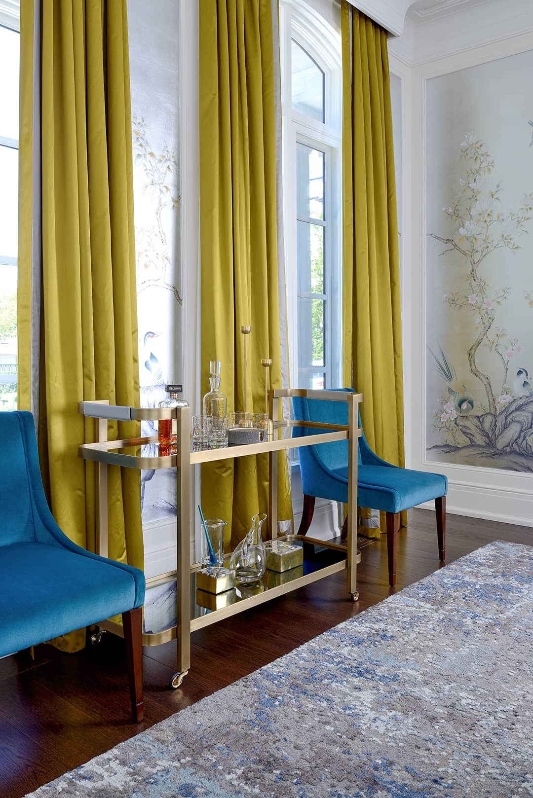 Opulent living room featuring bright gold drapes, teal velvet chairs, a brass bar cart with glass decanters, and a hand-painted wallpaper mural.