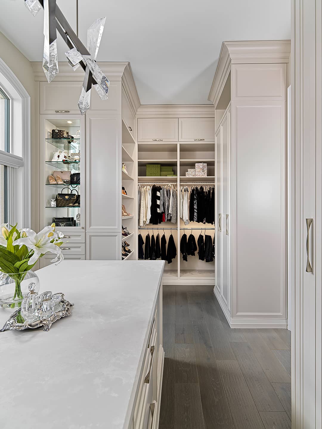 Luxury walk-in closet with custom cabinetry, glass shelving, center island, and elegant crystal chandelier.