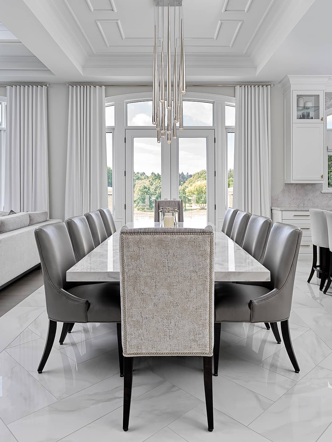 Modern dining room with marble table, upholstered chairs, tall pendant chandelier, and floor-to-ceiling windows with white drapery.