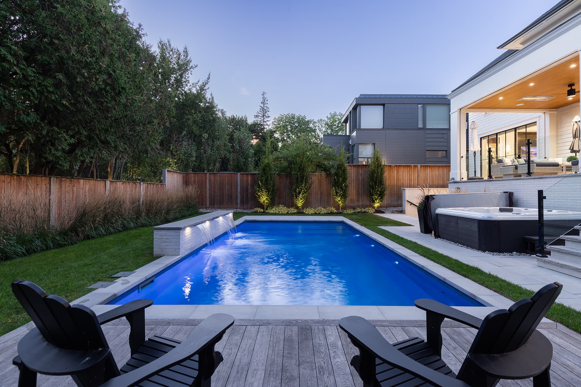 backyard with an illuminated swimming pool, integrated water feature, hot tub, landscaped lawn, and covered patio seating at dusk