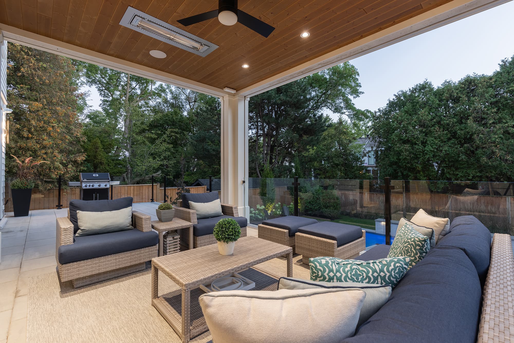 covered backyard patio with wicker lounge seating, wood ceiling with ceiling fan and heater, outdoor grill, and views of the landscaped yard and pool.