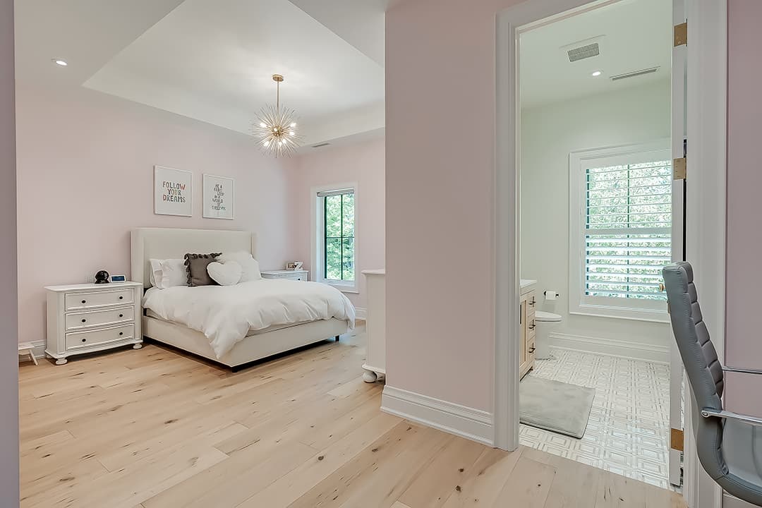 bedroom with soft pink walls, light wood floors, upholstered bed, brass starburst chandelier, and an adjacent bathroom.