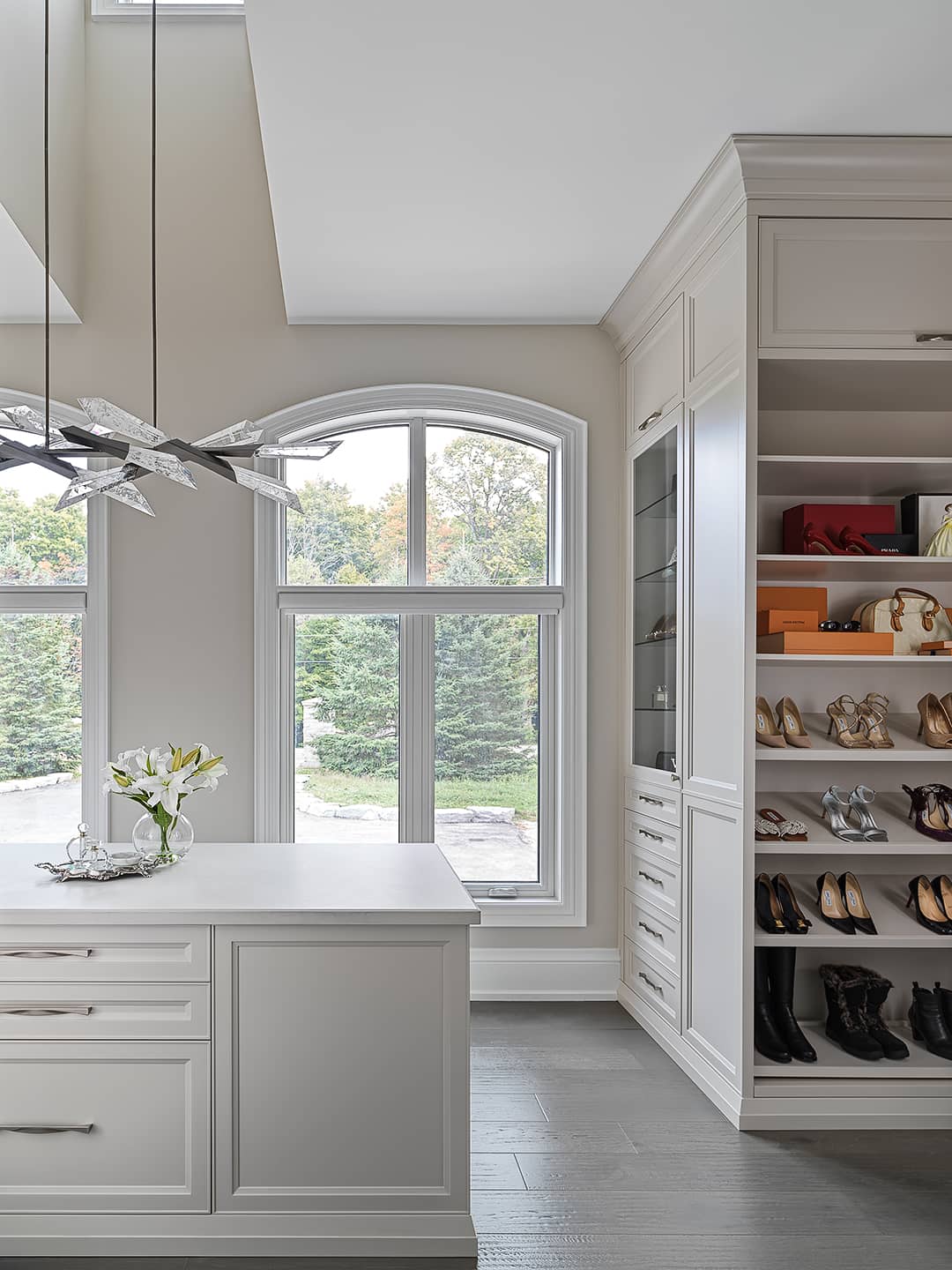 Bright walk-in closet with custom cabinetry, shoe display, large arched window, and modern crystal chandelier.