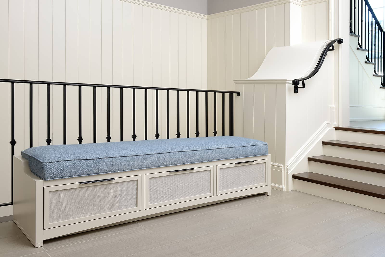 A built-in white bench with storage drawers and a light-blue cushion, set against white paneled walls and a black iron stair railing.