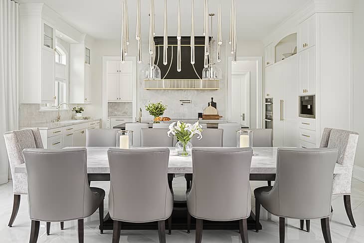 Bright modern kitchen and dining area with a marble-top table, grey upholstered chairs, and a statement brass and glass light fixture above a white cabinetry backdrop.