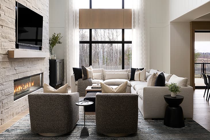 Modern chalet-style living room with a large sectional sofa, stone fireplace, and floor-to-ceiling windows overlooking a wooded landscape