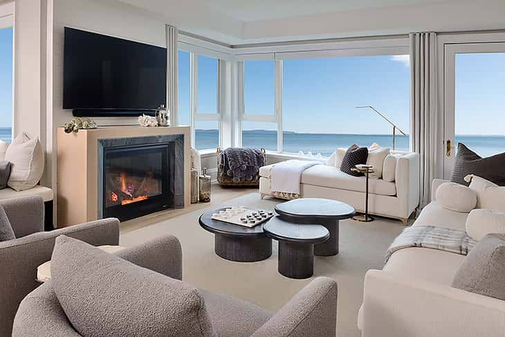 Coastal-inspired living room with neutral sofas, modern fireplace, round nesting coffee tables, and panoramic ocean views through large windows