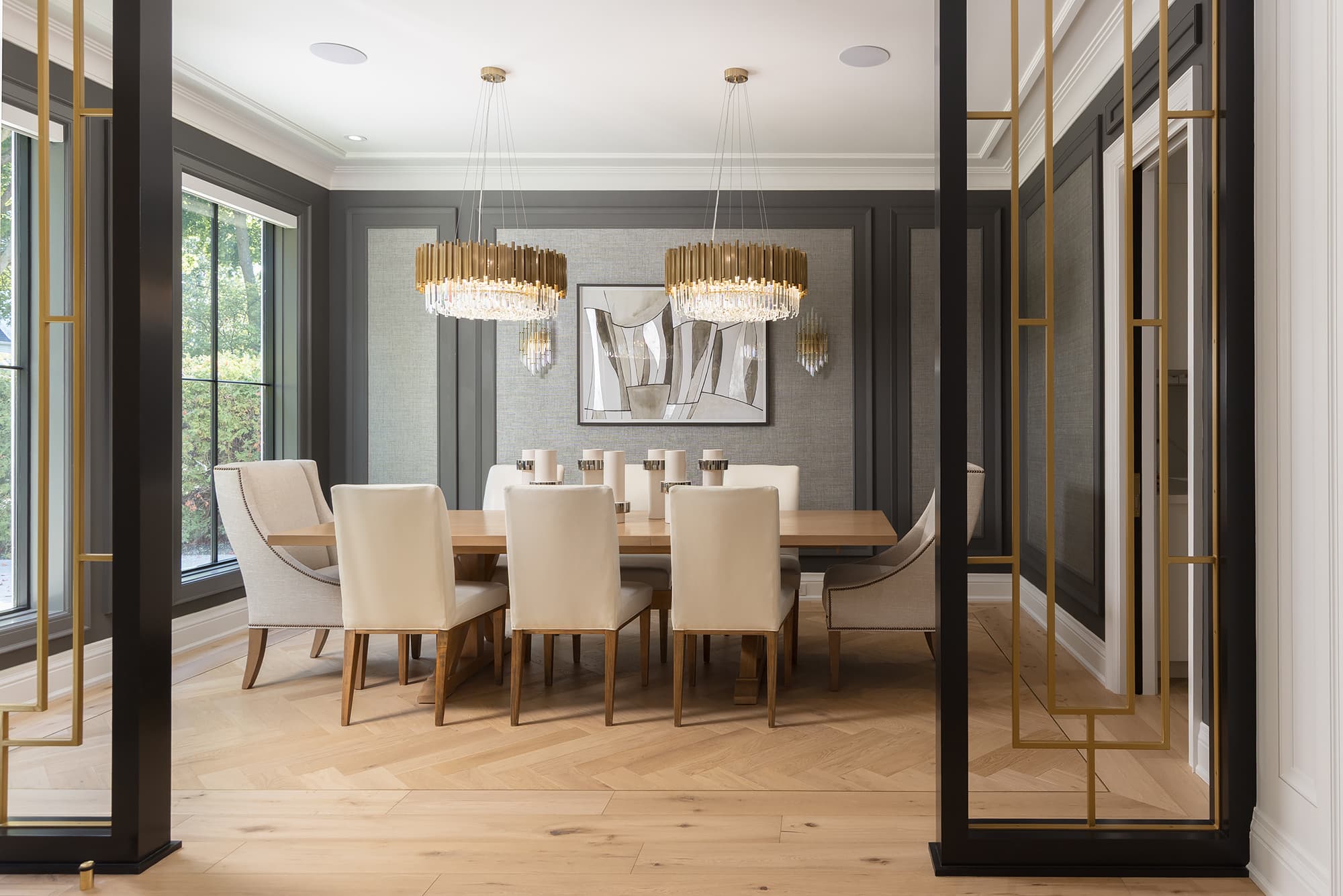Modern colonial dining room with light wood table, upholstered chairs, brass chandeliers, paneled gray walls, and black metal and brass room dividers.
