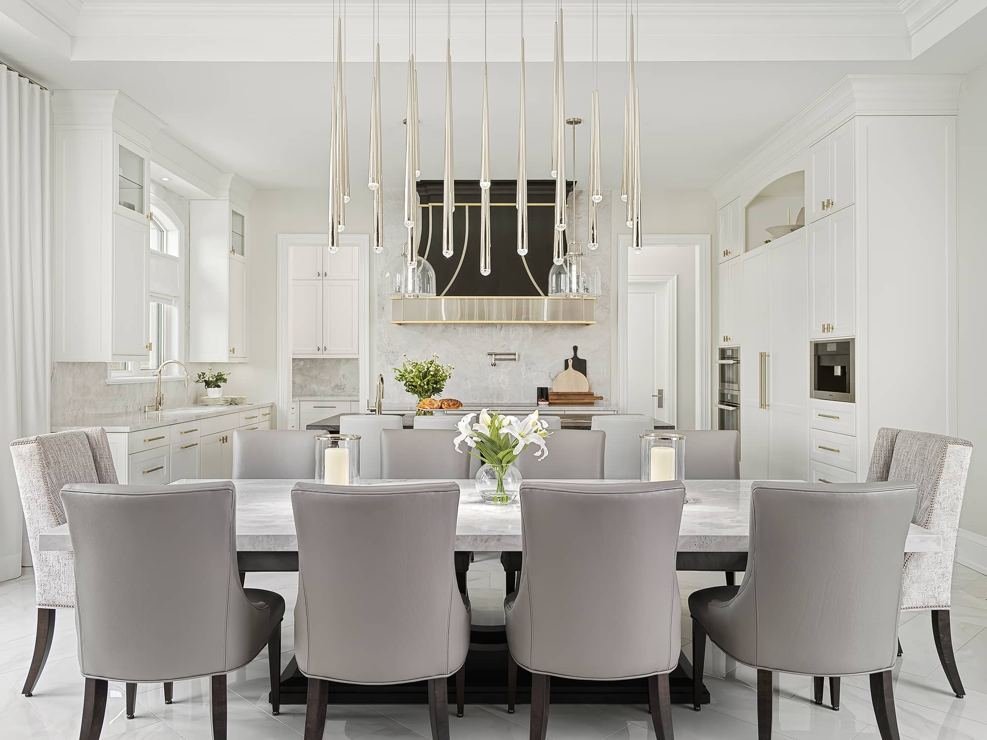 Elegant modern kitchen and dining area with marble table, custom black and gold range hood, white cabinetry, and contemporary pendant lighting, 10 chairs.
