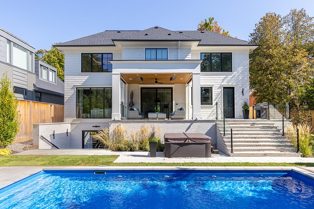 Rear exterior of a modern colonial home with white siding, covered patio, black-framed windows, stone steps, landscaped yard, and an in-ground swimming pool.