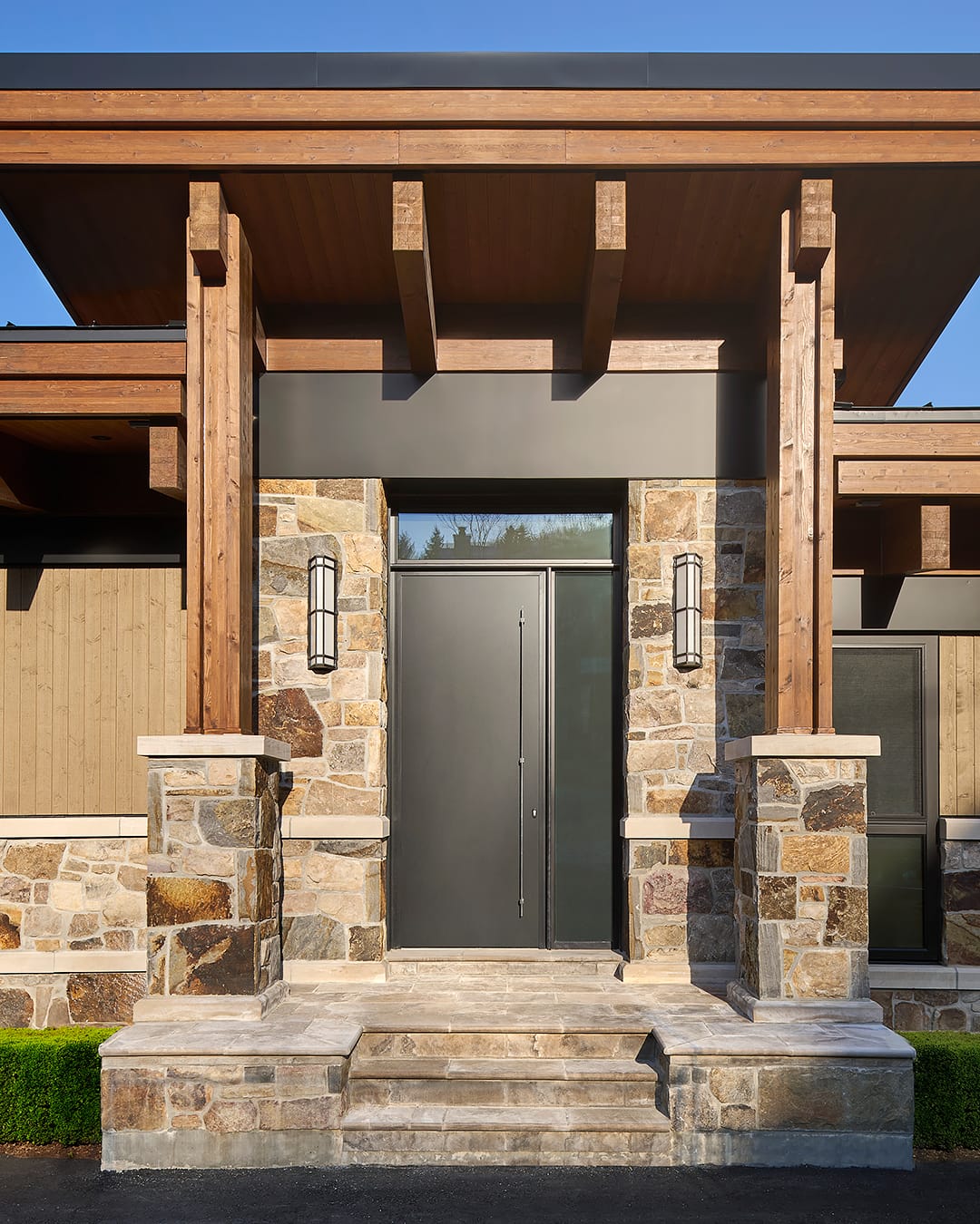 Exterior front entrance of a modern luxury chalet featuring a custom oversized black door, natural stone cladding, and exposed timber columns with contemporary sconce lighting