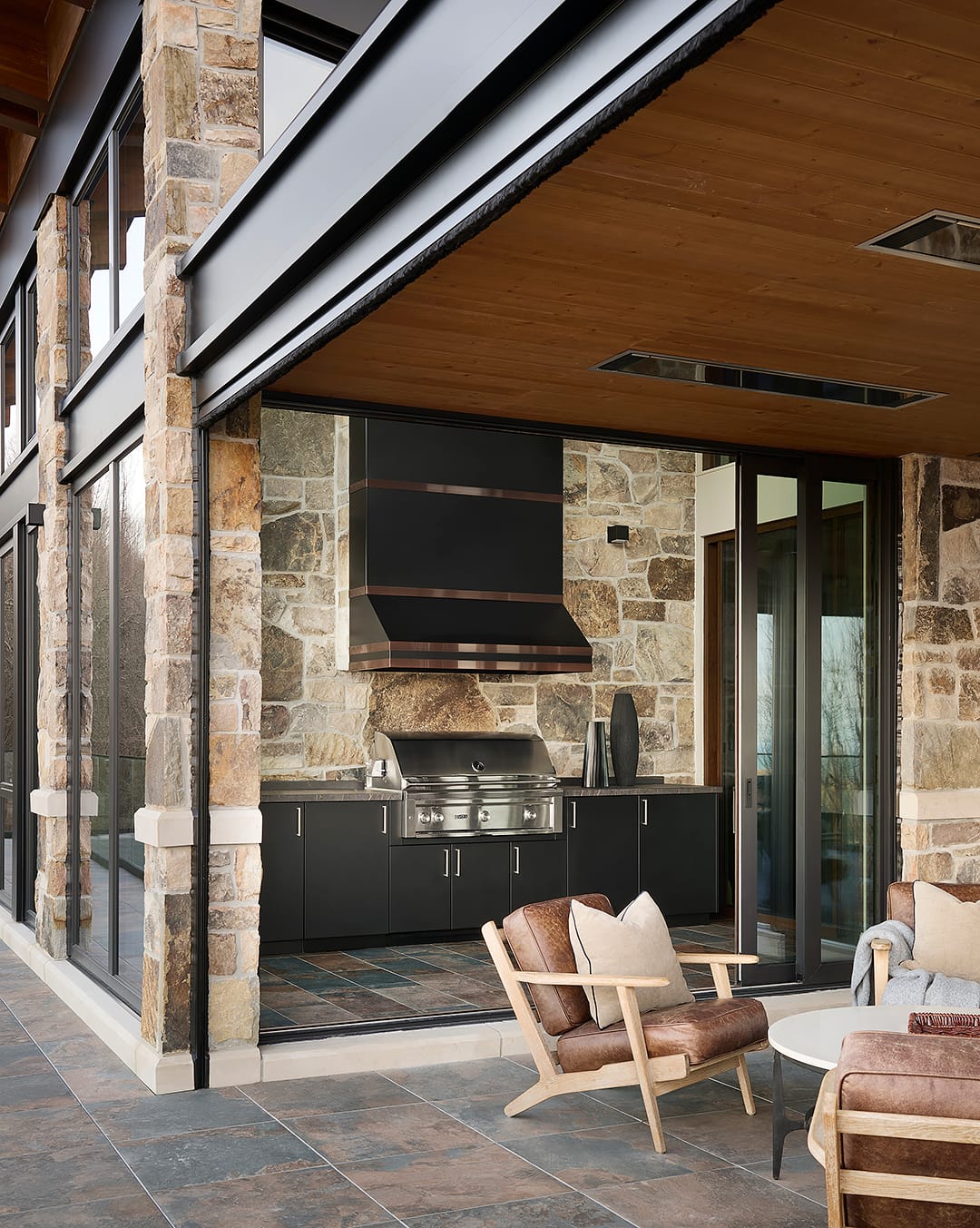 A modern luxury indoor grilling room featuring a professional grade stainless steel grill with a custom black and copper oversized vent hood set against a natural stone accent wall.