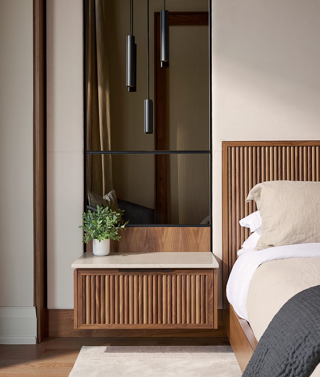 A close up of a custom walnut fluted floating nightstand with a stone countertop and integrated smoked mirror wall panel featuring black pendant lights in a luxury bedroom