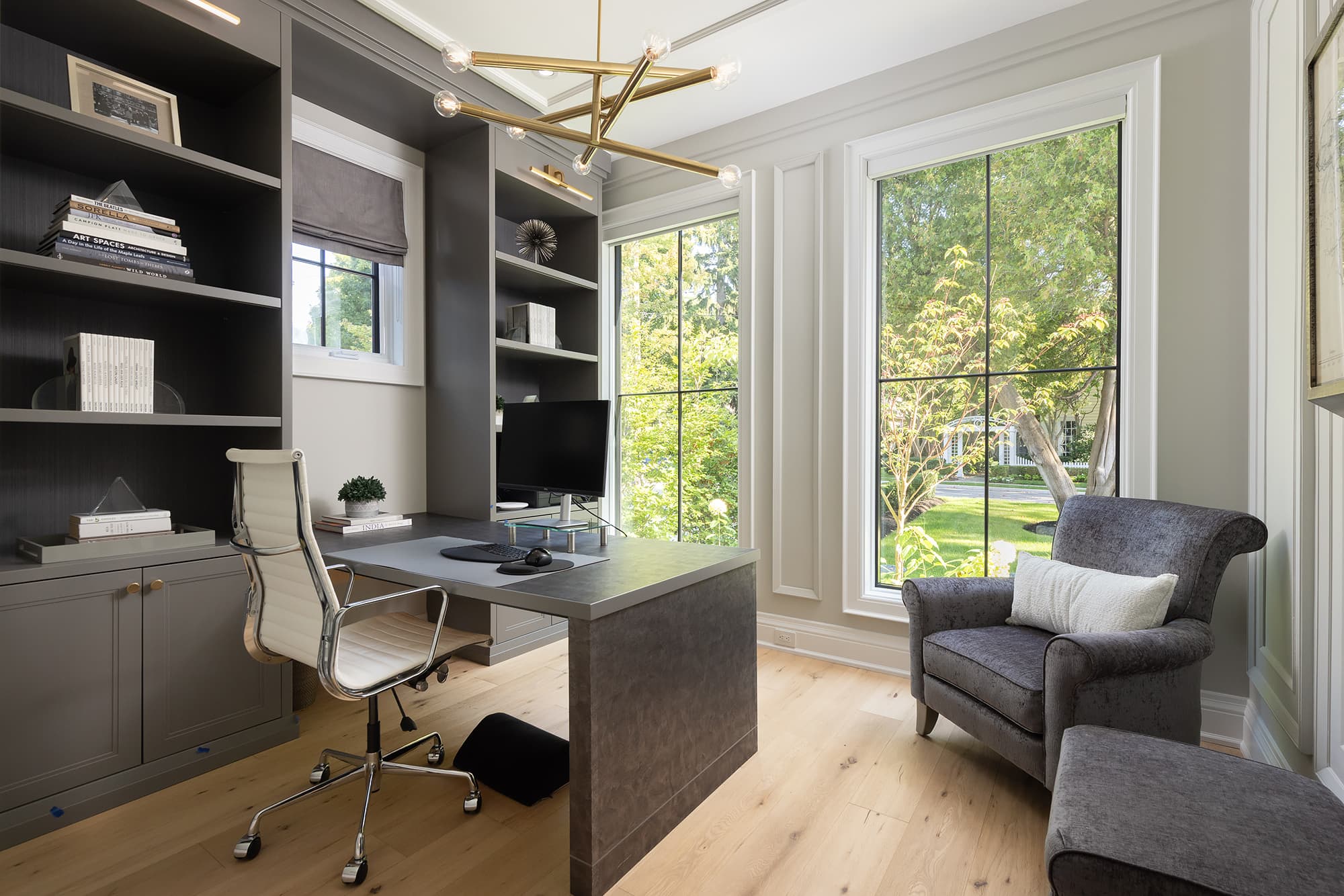 home office with built-in shelving, minimalist desk, brass chandelier, large windows, and a cozy reading chair overlooking the front yard