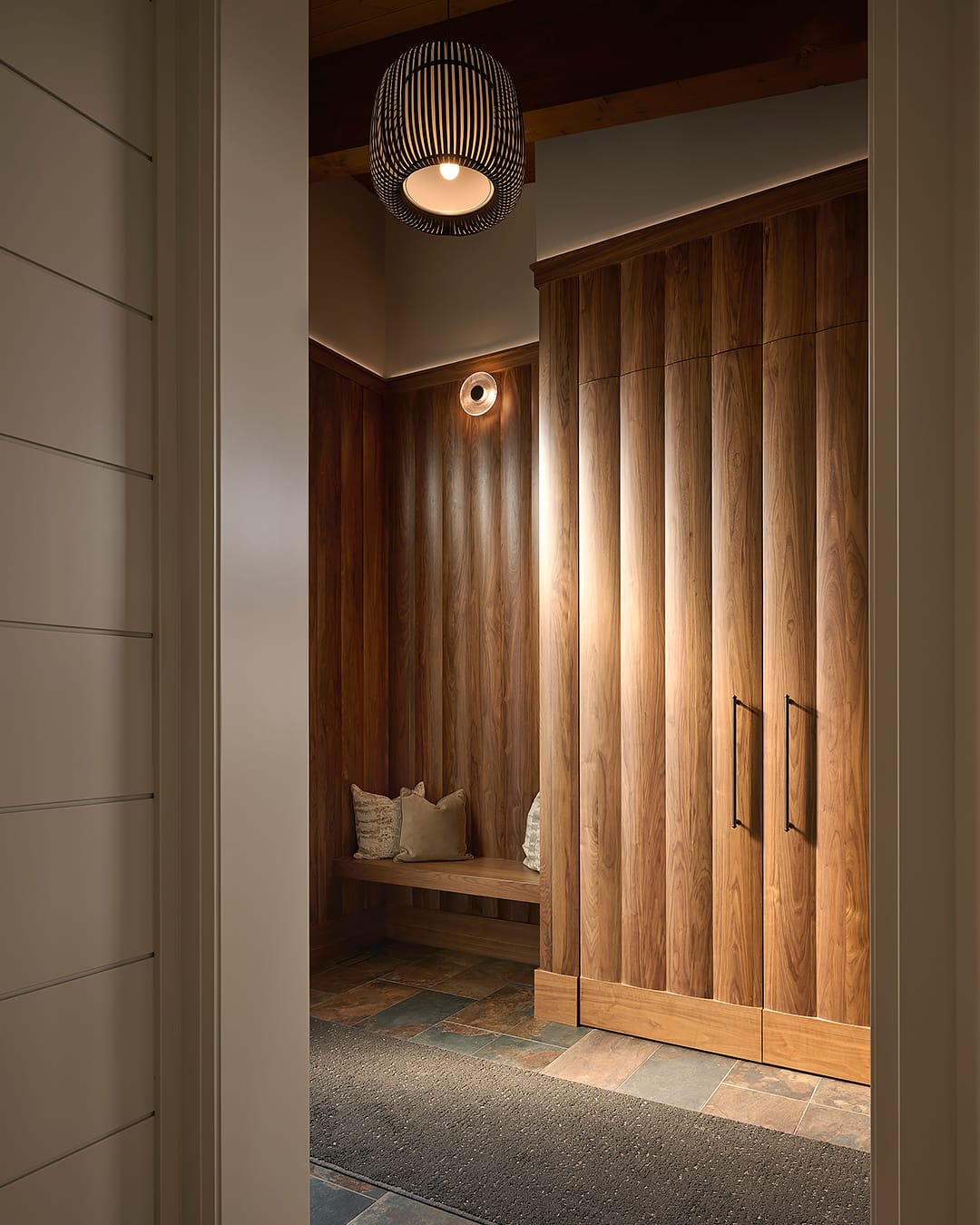 A luxury mudroom hallway featuring floor to ceiling custom walnut wood paneling with integrated storage closets, a built-in wood bench, and a modern black slatted pendant light.