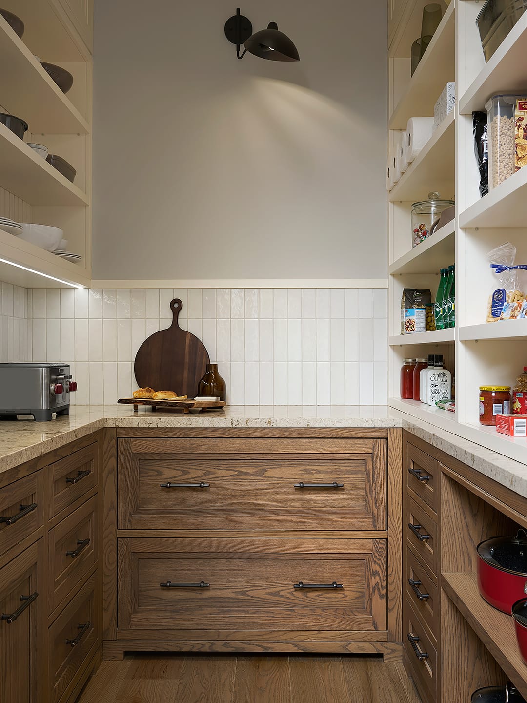 A luxury walk-in pantry featuring custom light oak cabinetry with matte black pulls, granite countertops, and a cream zellige tile backsplash with integrated under-cabinet lighting