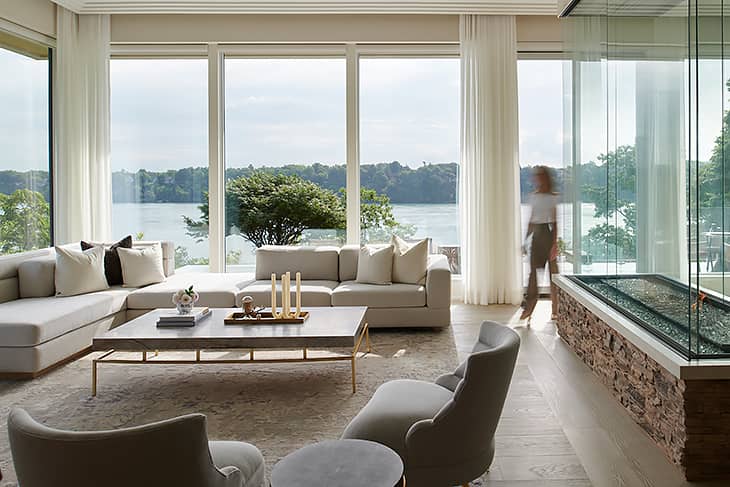 Modern waterfront living room with a light neutral sectional, large floor-to-ceiling windows, and views overlooking the lake.
