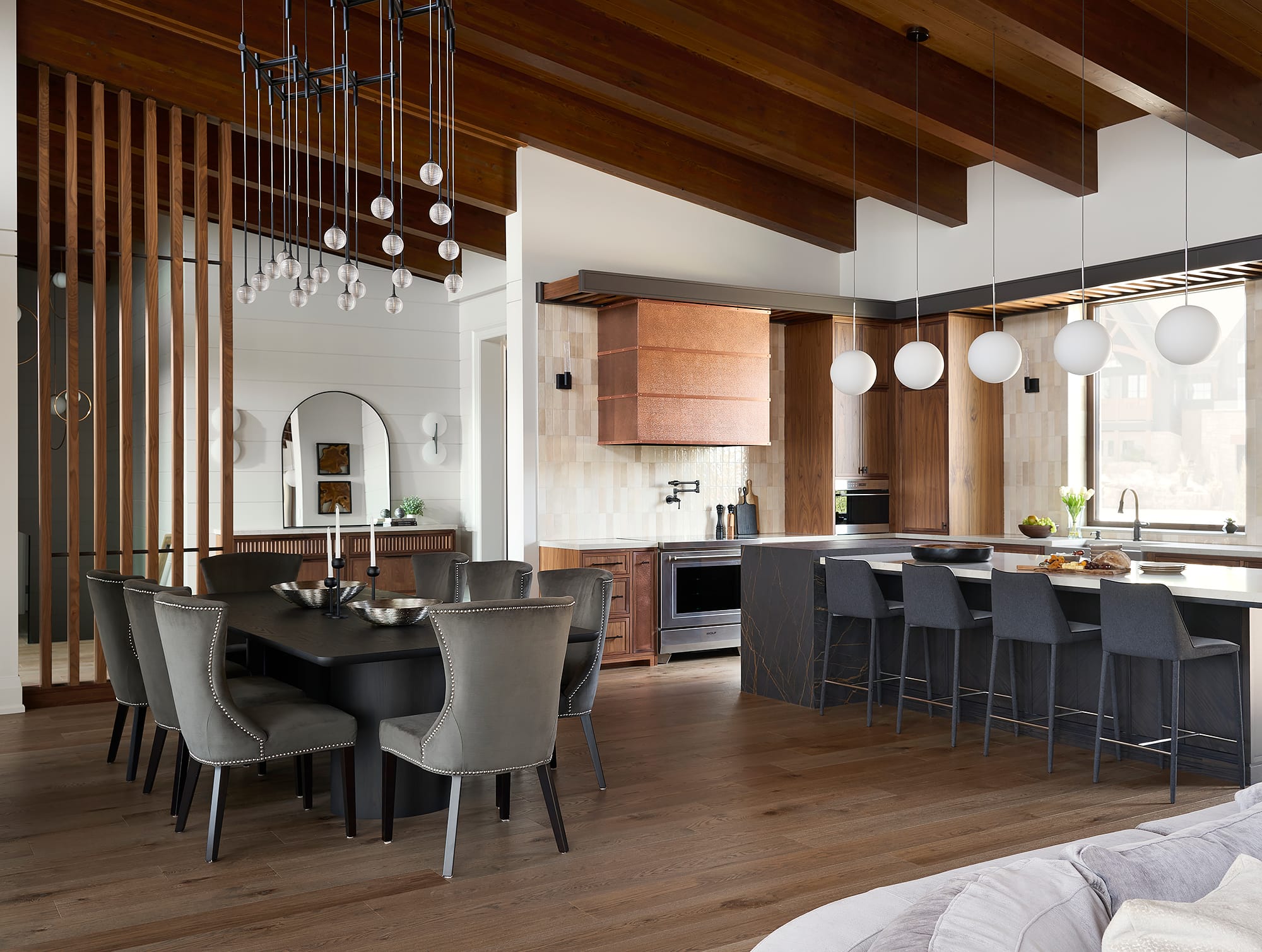 An open-concept dining area featuring a dark wood table, grey upholstered chairs, and a modern statement chandelier under exposed wood beams, leading into the luxury kitchen