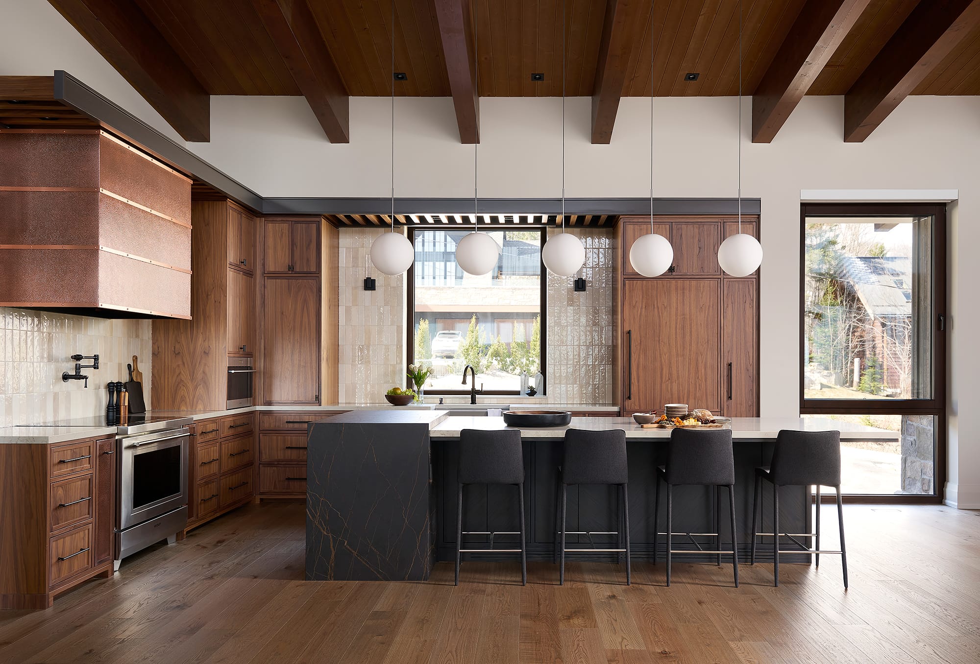 A modern luxury kitchen in a Collingwood ski chalet featuring walnut cabinetry, a custom copper range hood, wood-beamed ceilings, and a large black waterfall island with globe pendant lighting.