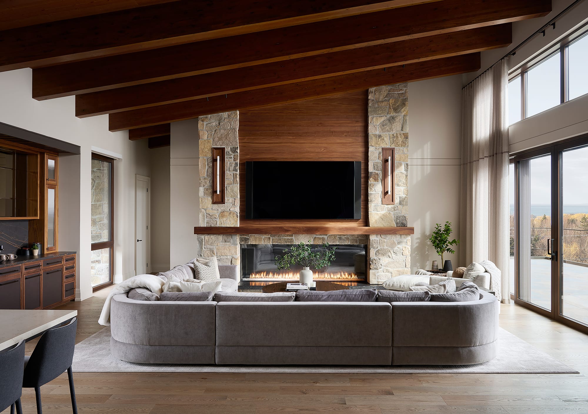 A modern living room in a Collingwood ski chalet featuring a large stone fireplace with walnut panels, a curved grey sectional sofa, and vaulted wood-beamed ceilings