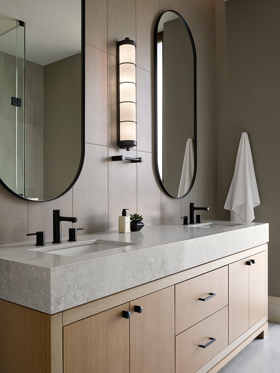 Modern luxury bathroom featuring a light oak double vanity with a thick stone countertop, matte black faucets, and two large oval mirrors separated by a contemporary black and glass wall sconce