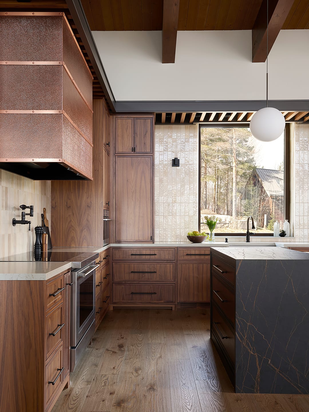 Luxury kitchen in a modern chalet featuring custom walnut cabinetry, an oversized hammered copper vent hood, and a dark marble waterfall island set against cream zellige wall tile