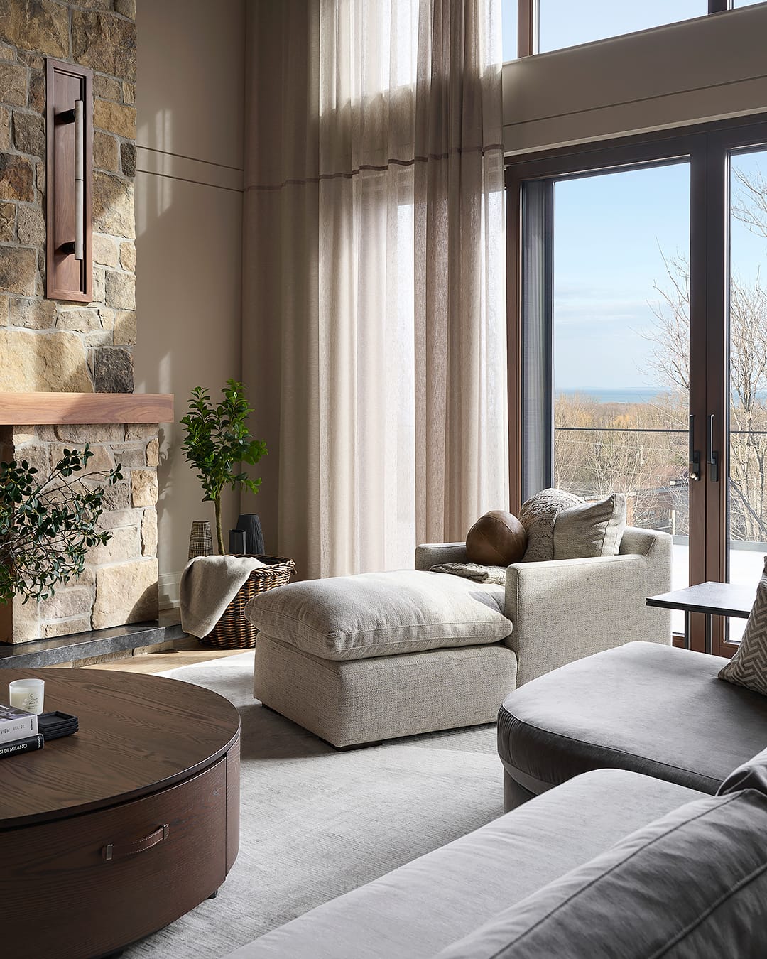 Luxury living room in a modern alpine chalet featuring a floor-to-ceiling stone fireplace, a cozy neutral chaise lounge, and massive windows with floor-to-ceiling sheer drapery overlooking a winter landscape