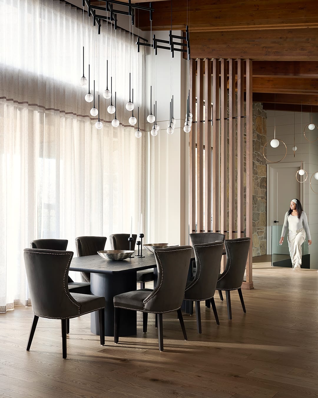 Luxury dining room in a modern chalet featuring an oversized black pedestal table, velvet studded chairs, a multi-globe tiered chandelier, and a floor-to-ceiling oak slat room divider