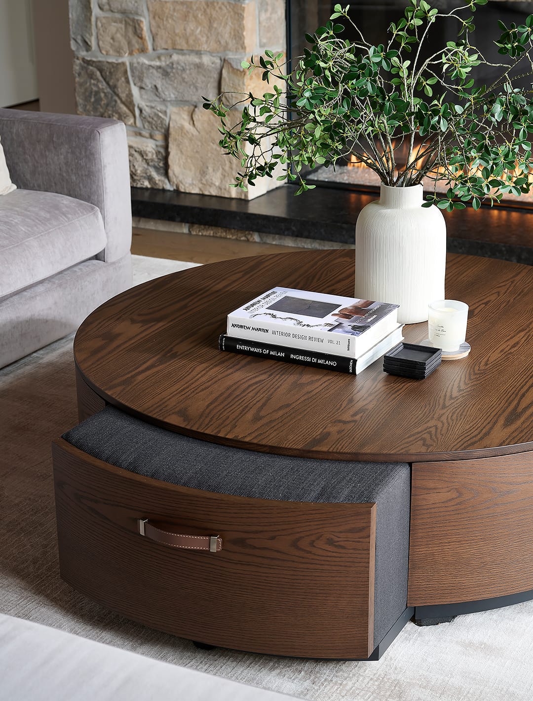 A close up of a custom circular walnut wood coffee table with a pull-out upholstered storage ottoman featuring a leather handle in a luxury living room