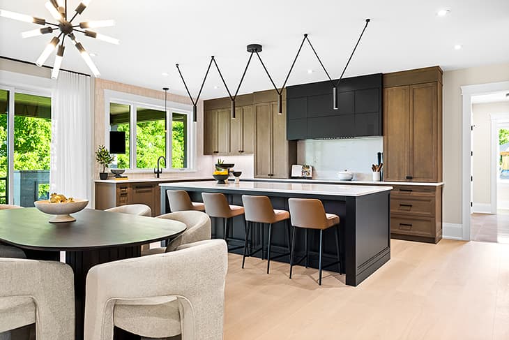Modern traditional kitchen with two-tone wood and black cabinetry, a large island with bar seating, sculptural pendant lighting, and an open dining area filled with natural light.