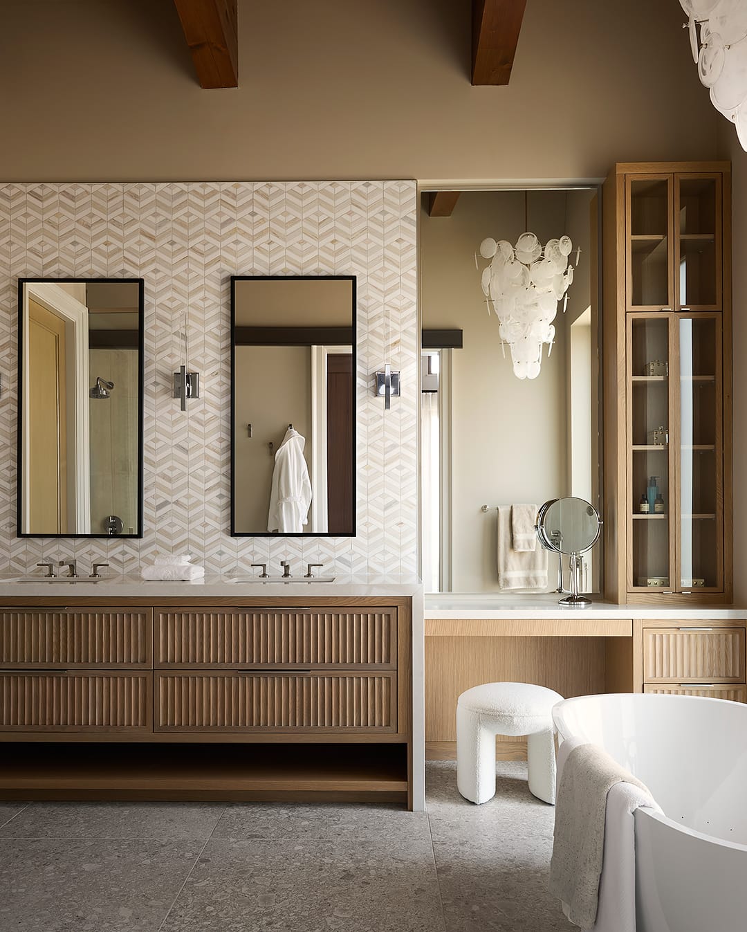 Luxury primary ensuite featuring a light wood fluted double vanity, geometric marble tile backsplash, and a dedicated makeup station with a built-in linen tower and capiz shell chandelier