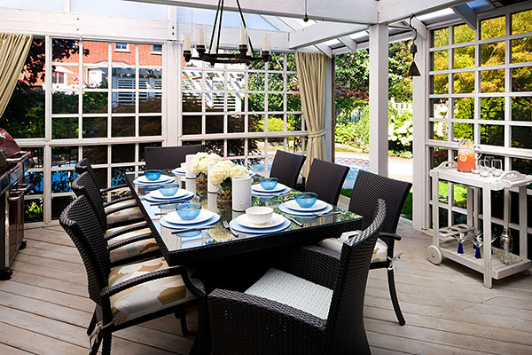 Covered outdoor dining room with wicker chairs, a glass-top table set for eight, and garden views through grid-style windows.