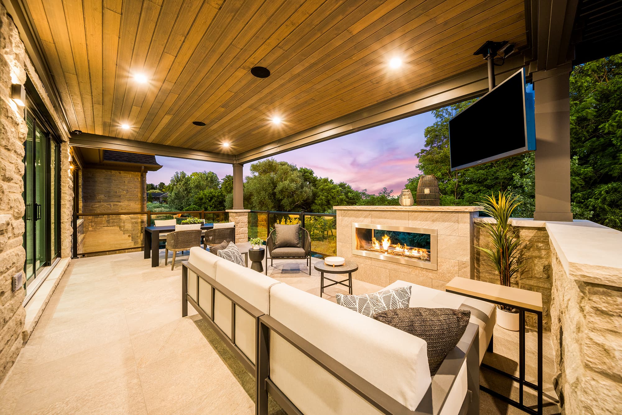 covered outdoor terrace with a stone fireplace, comfortable seating, wood ceiling, integrated lighting, and views of the surrounding landscape at dusk