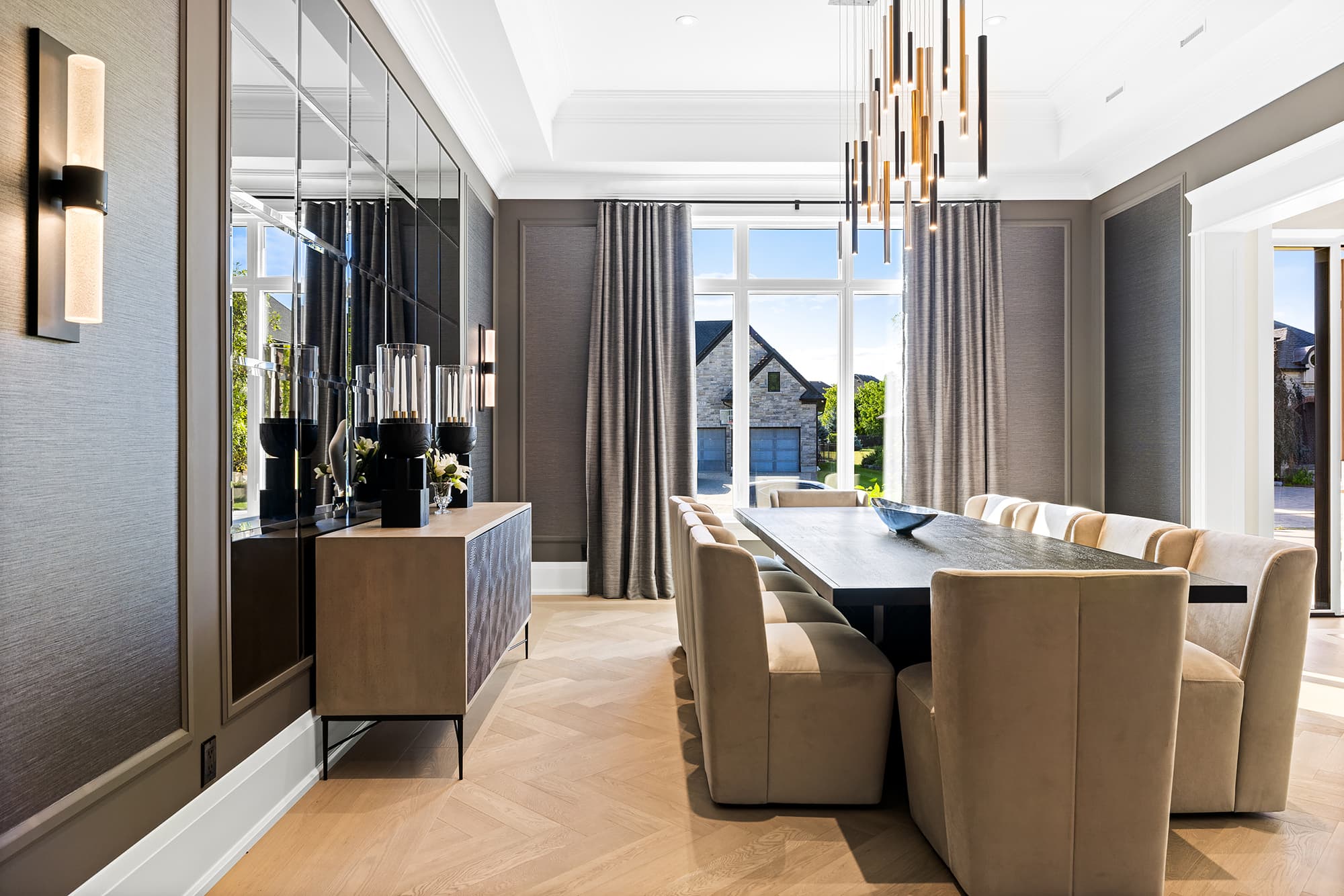 Traditional dining room with a long wood dining table, upholstered seating, custom pendant lighting, mirrored accent wall, and soft natural light from large windows.