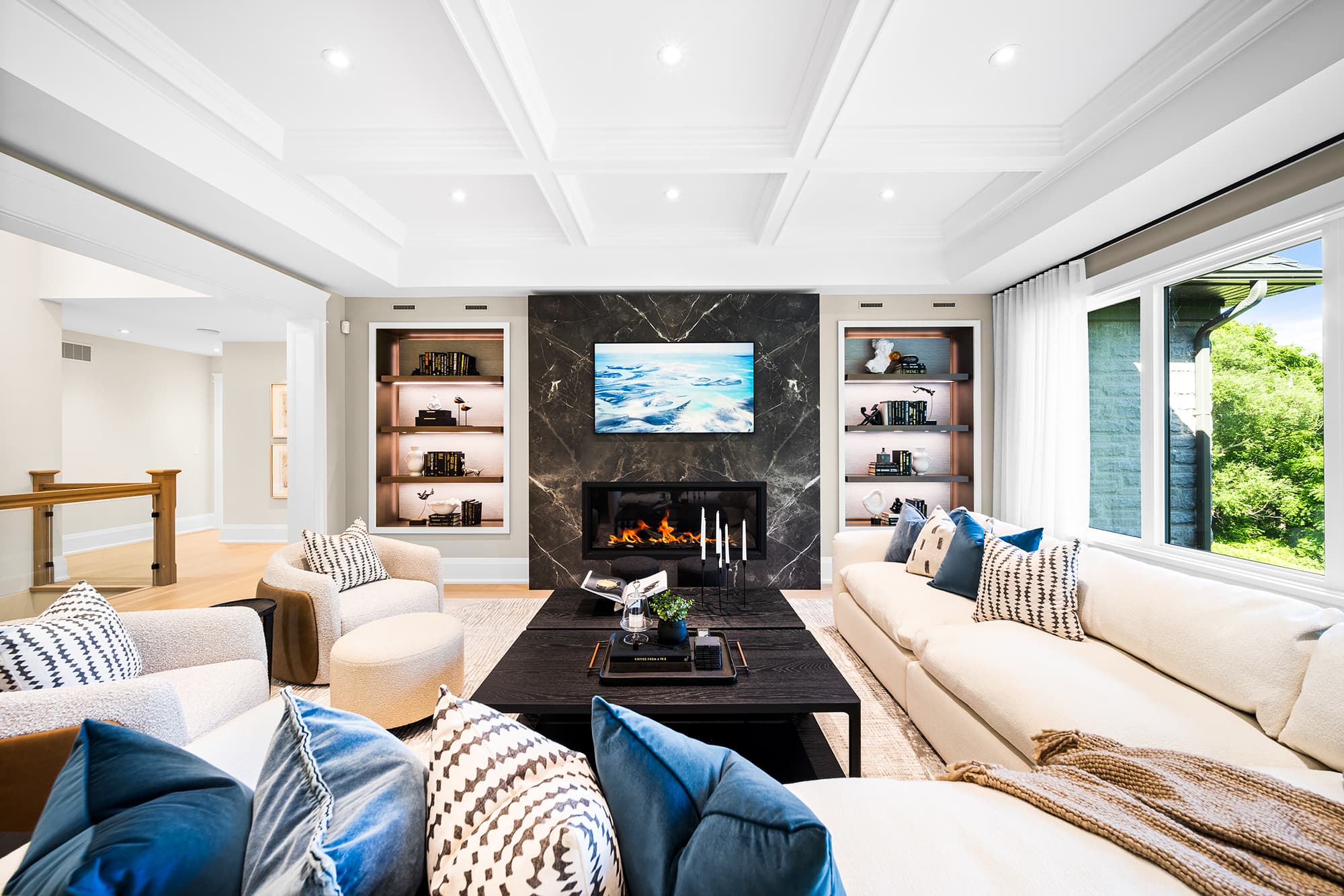 Living room with a slab fireplace, custom built-in shelving, coffered ceiling, and layered seating arranged around a central coffee table.