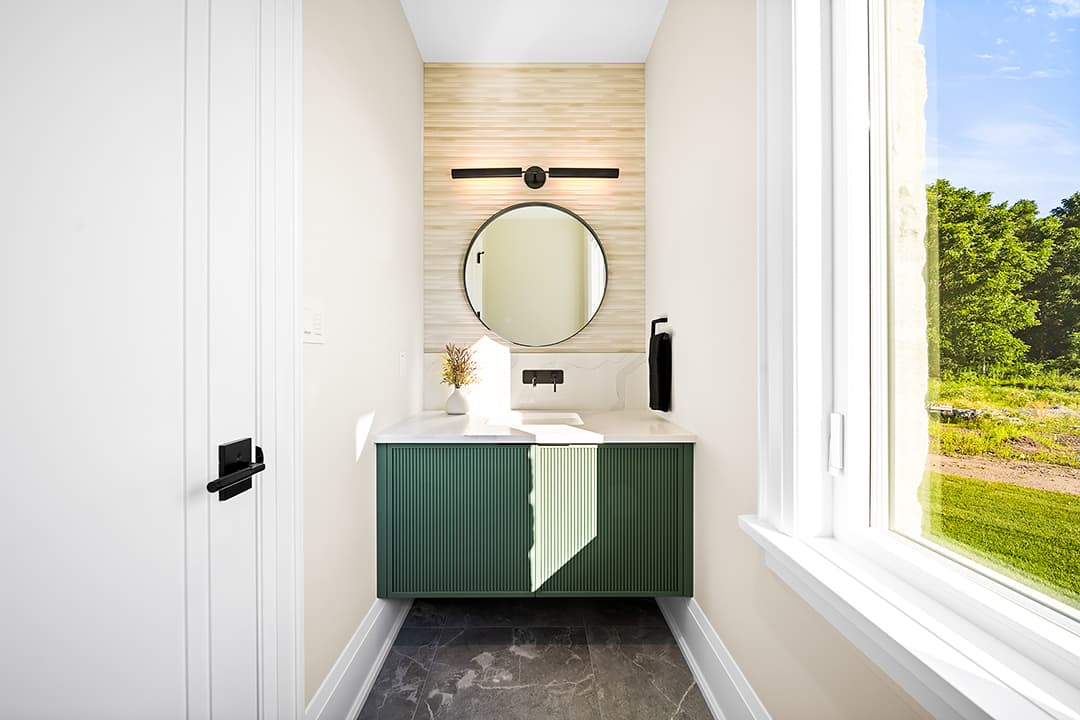 Powder room with a custom fluted vanity, stone countertop, round mirror, textured wall detail, and natural light from a large window.