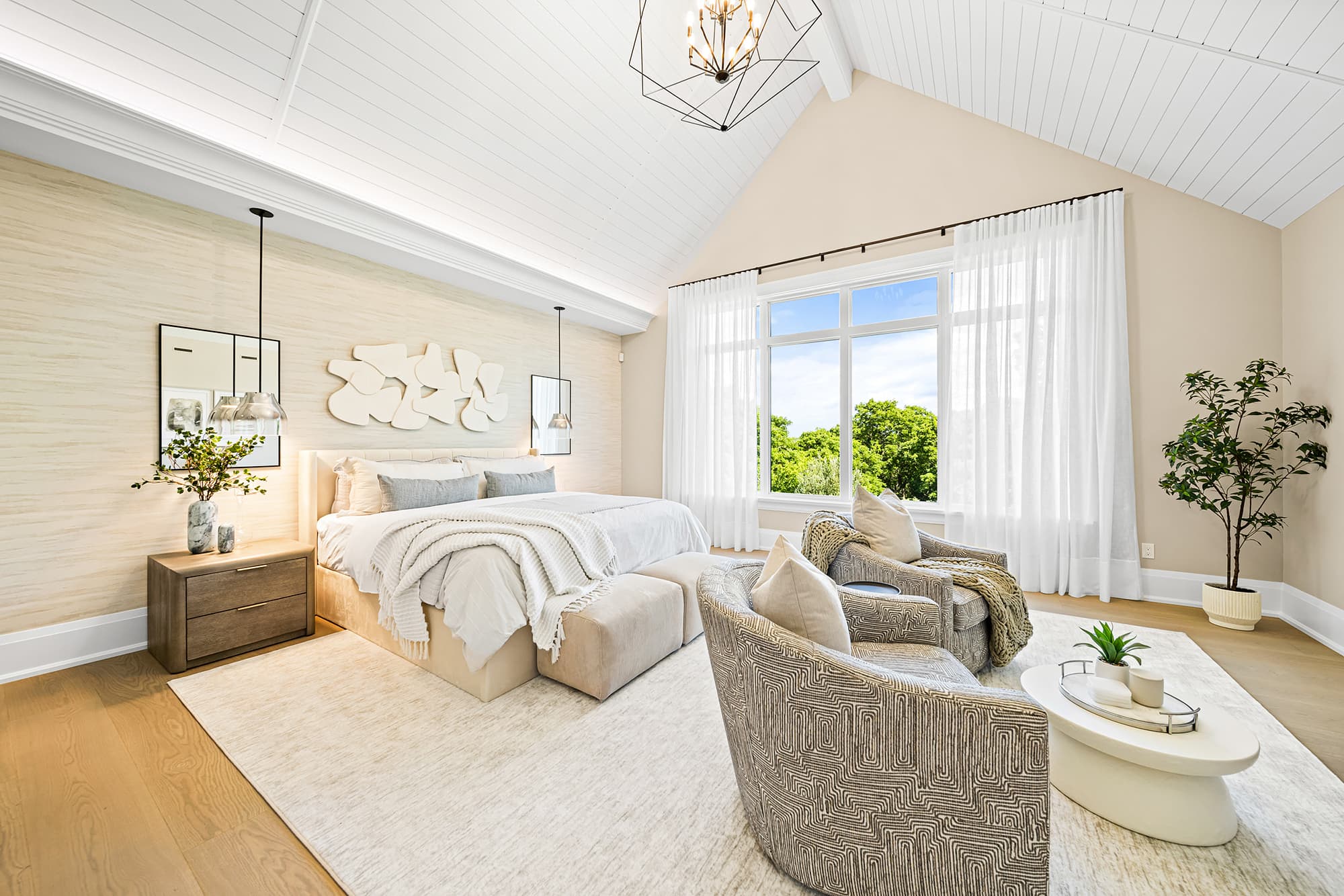 Traditional primary bedroom with a vaulted shiplap ceiling, upholstered bed, sitting area with lounge chairs, and large windows dressed in sheer curtains.