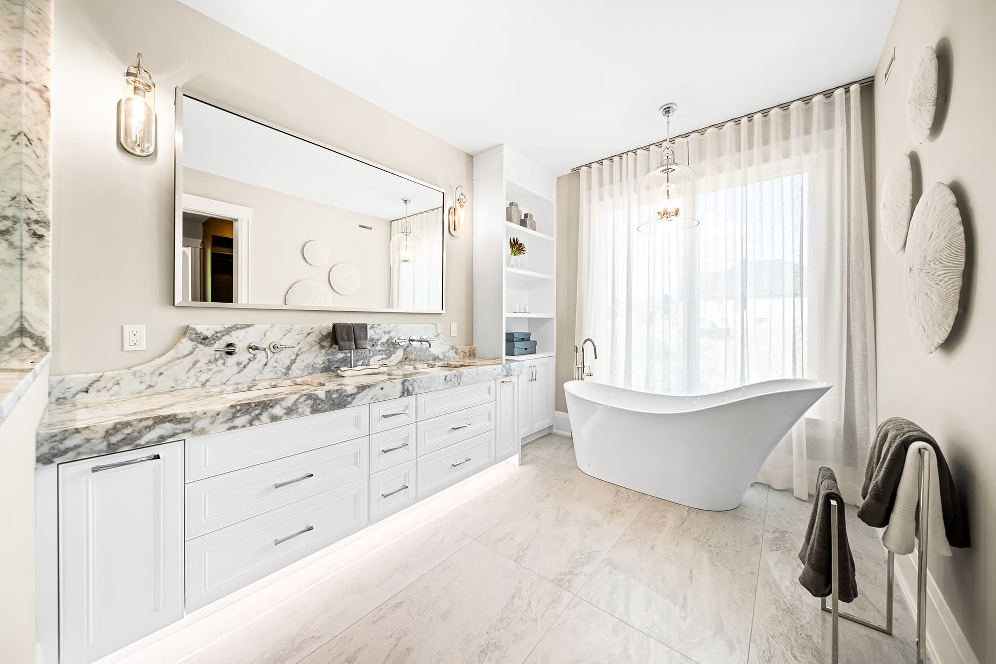 Primary ensuite bathroom with a marble vanity, freestanding soaking tub, soft neutral finishes, and natural light filtered through sheer curtains.
